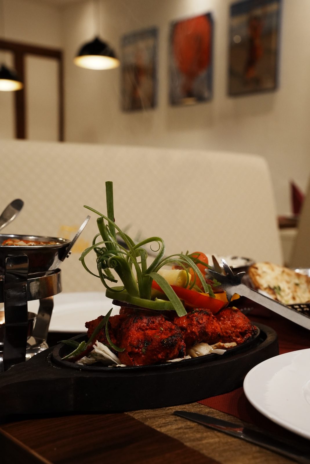 Tandoori-Hähnchen mit Paprika und Frühlingszwiebeln auf der heißen Platte, dahinter die bestickte Sitzbank und Hängelampen im Gastraum